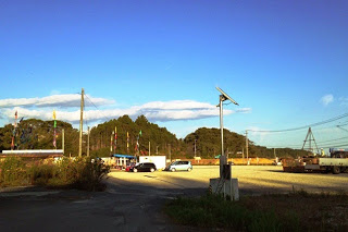 青空の下、旗が立ち並ぶ工事現場の入口付近に数台の車が止まっており、周囲に山や空き地が広がる様子の写真