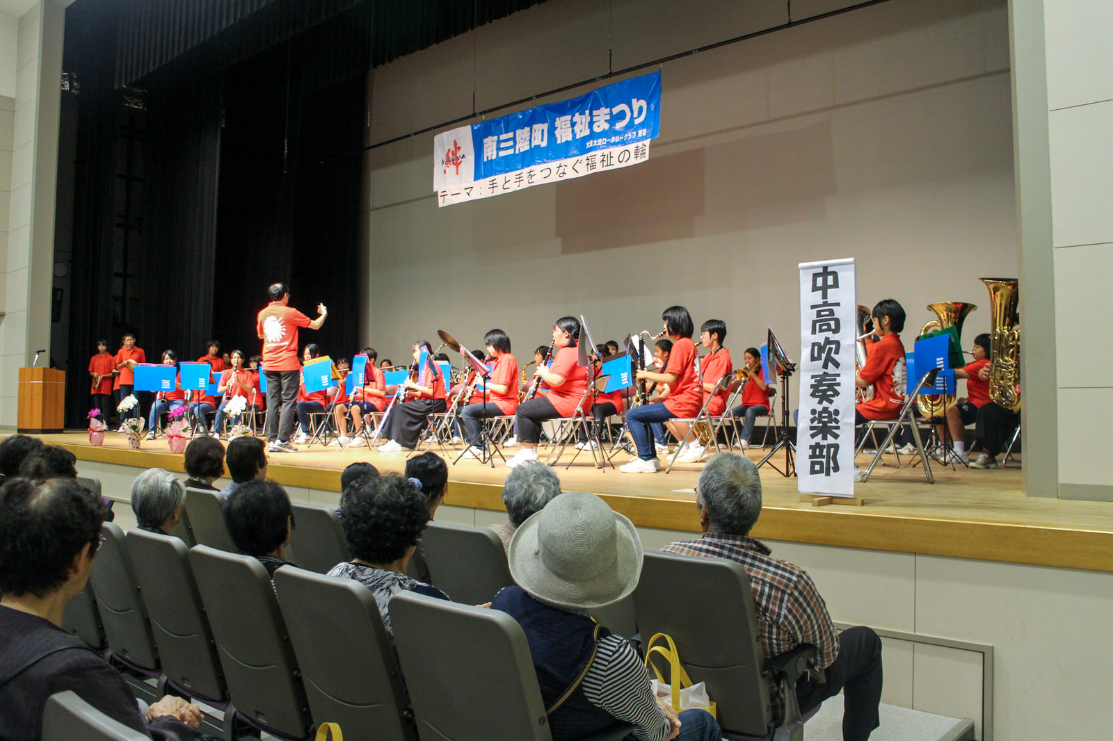 「南三陸町福祉まつり」と書かれたステージ上で、観客席でお客さんが多数座って楽しんでいるなか、指揮者が前に立って指示を出し、お揃いの赤いTシャツを着た中高生の吹奏楽部が演奏している様子の写真