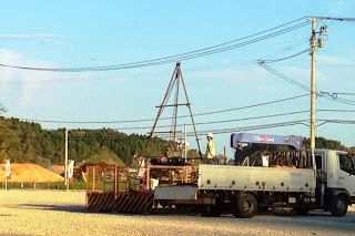 広い空き地にクレーン付きのトラックが停まり、作業員が作業をしている建設現場の写真