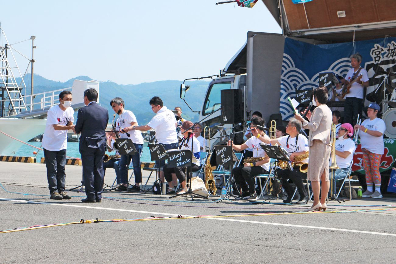 メンバーが「Liberty of Music」の譜面台の前に座っている屋外のステージ前で、白いシャツを着た代表の男性とスーツ姿の佐藤町長が握手している様子の写真