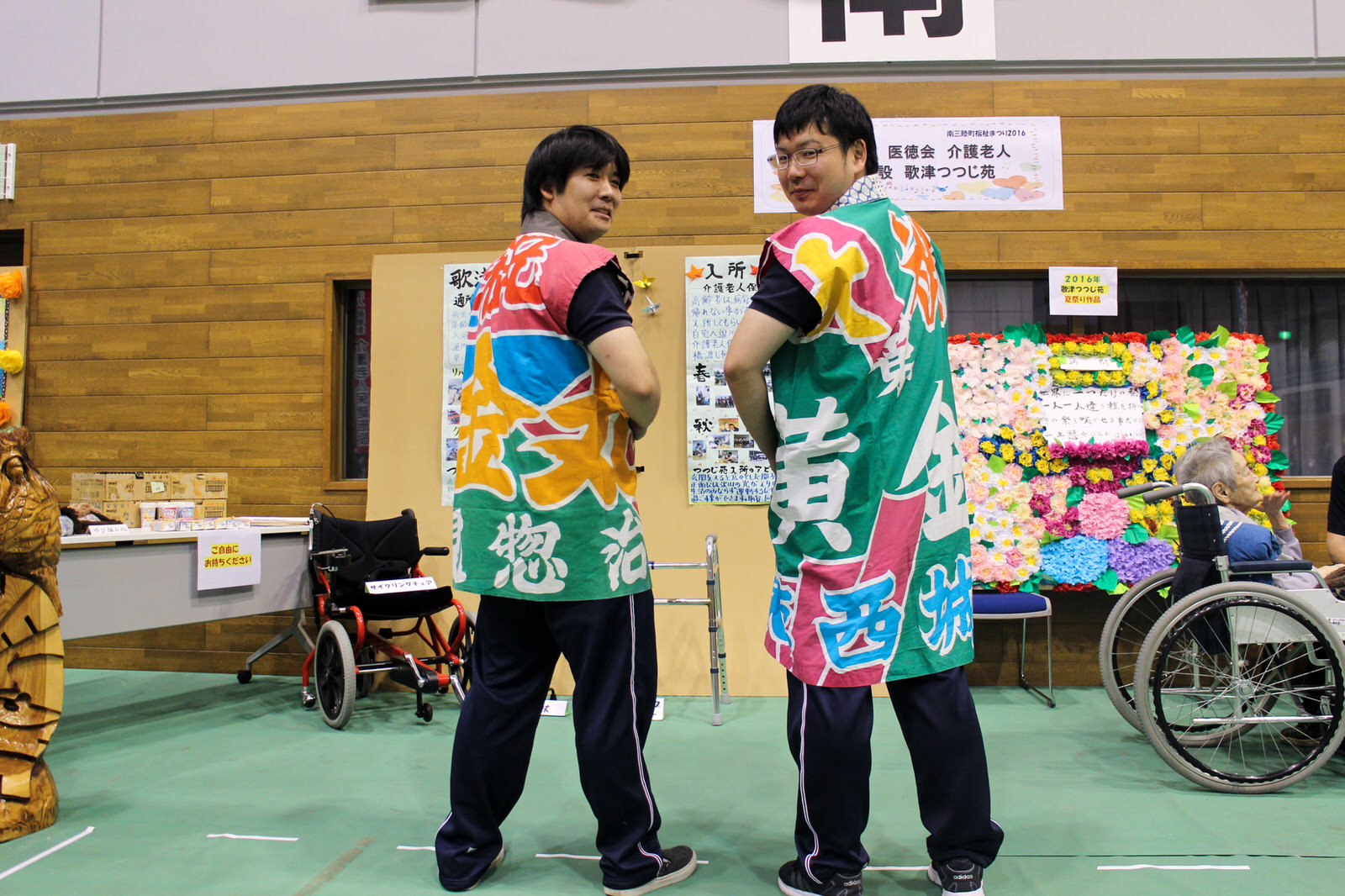 背景には花で飾られた掲示物や車いすの高齢者がおり、カラフルな文字が大きく書かれた大漁旗で作られた法被を着た二人の男性が、背中をこちらに向けて笑顔でポーズを取って並んでいる、介護施設の紹介ブース前での記念撮影の写真