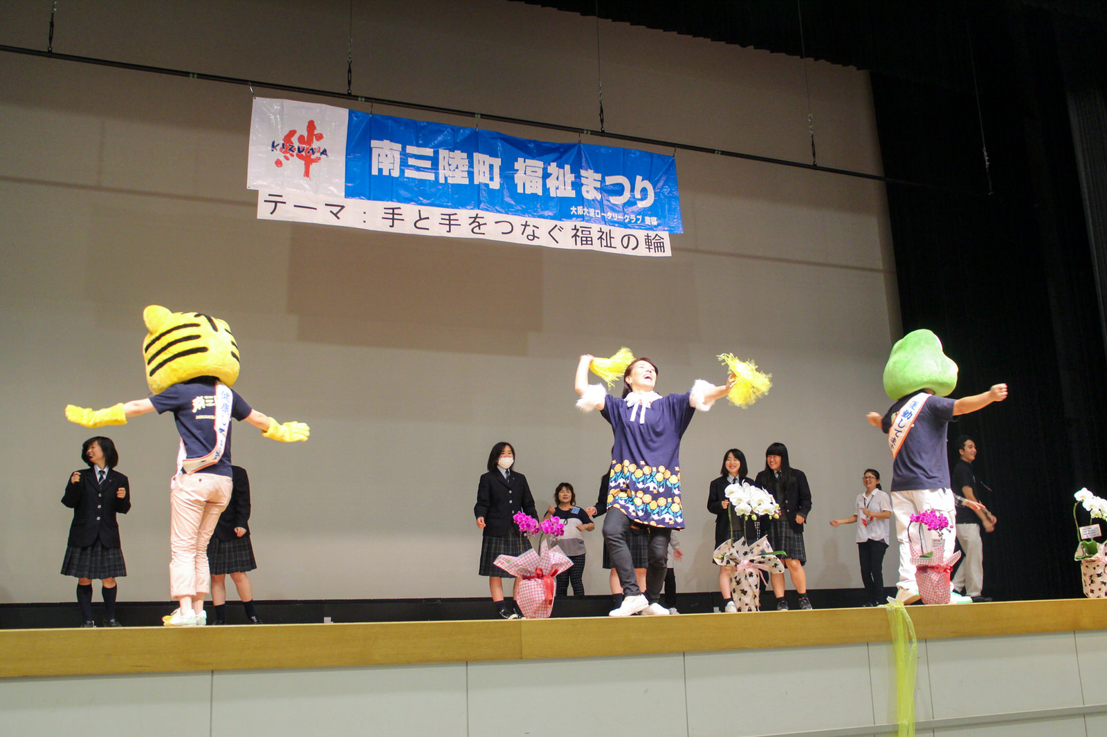 「南三陸町福祉まつり」と書かれた横断幕が掲げられたステージ上で、後ろには黒い制服を着た女子学生たちや女性が間隔をあけて立ち、中央に黄色のボンボンを両手で持ち踊っている女性と、両隣の黄色いトラの被り物と緑色のカエルの被り物をかぶった2人が手を広げて横を向き踊っている写真