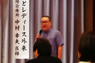 背景の垂れ幕に「レディース外来 南三陸診療所 中村幸夫医師」と書かれている前で、青色のシャツを着た男性がマイクを持って話している写真