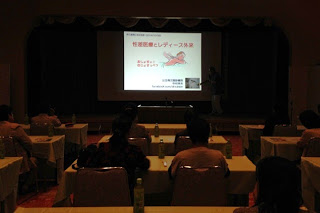 薄暗い講演会場で、前方に設置された資料が映し出されたスクリーンを数十人の参加者が見ている写真