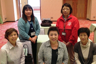カーペット敷きの会場内で、職員証や赤いジャンパーを着用している2人の女性と3人が笑顔で並んでいる記念写真