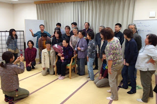 白いカーテンの前で大勢の参加者が集まり、花束を手に持ち椅子に座った女性を中心に並び記念撮影を行おうとしている写真