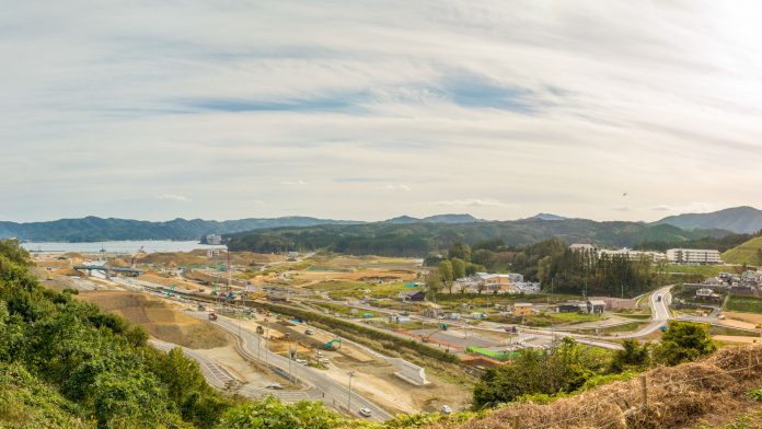 敷地内にはさまざまな建設機械、車両、資材が広く配置されており、複数の道路や通路が確認でき一部のエリアでは開発や掘削が進行中の様子が見られ、背景には緑に覆われた丘や山々が広がり、空は部分的に雲がかかっている写真