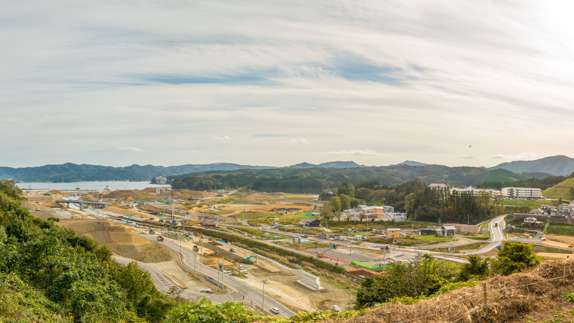 敷地内にはさまざまな建設機械、車両、資材が広く配置されており、複数の道路や通路が確認でき一部のエリアでは開発や掘削が進行中の様子が見られ、背景には緑に覆われた丘や山々が広がり、空は部分的に雲がかかっている写真