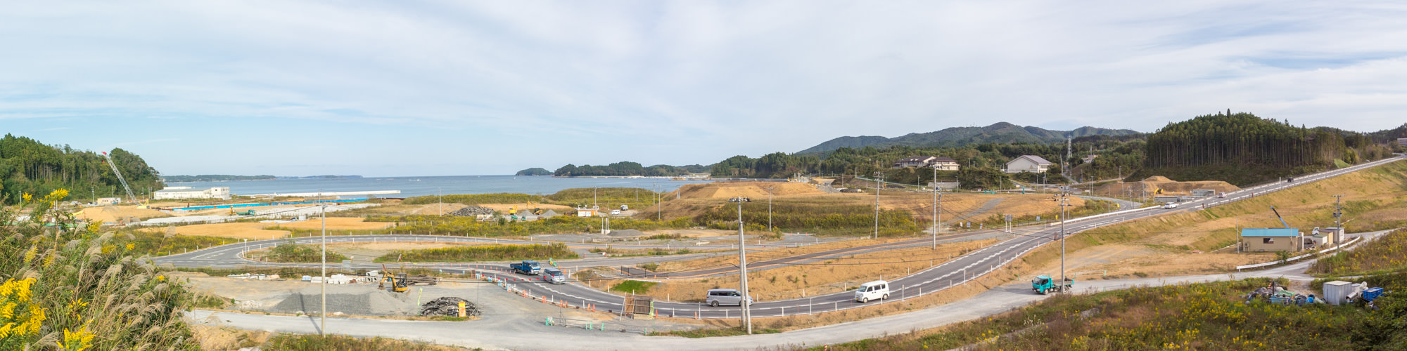 複数の道路が敷かれており、建設車両や機材が点在し背景には海が広がり、いくつかの島と森林に覆われた丘が見えている写真