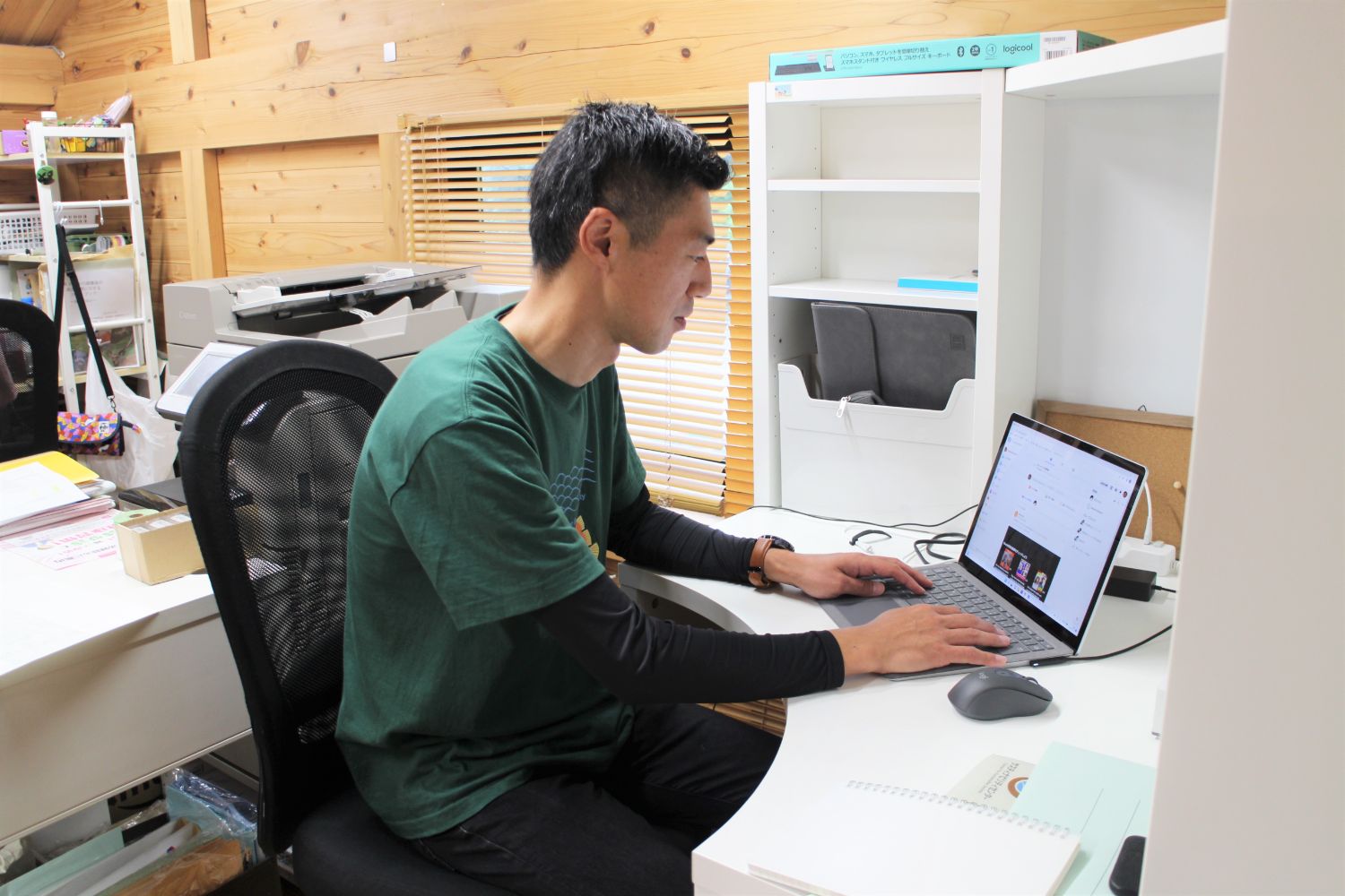 緑色のTシャツを着た小林さんが、白いデスクでノートパソコンを操作しながら仕事をしている様子の写真
