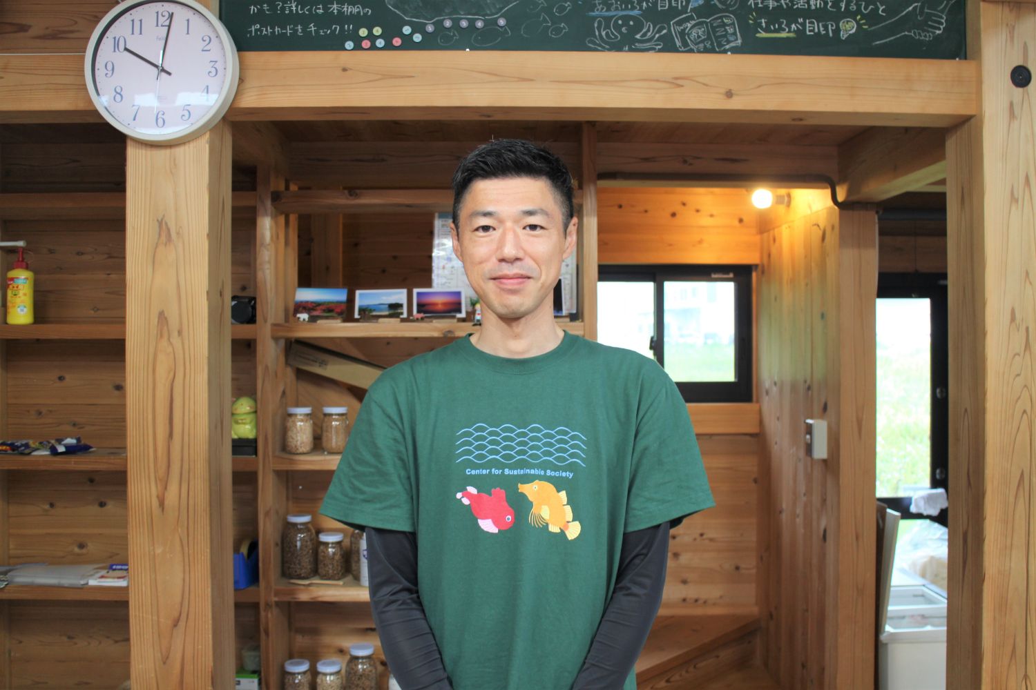 木造の室内で、緑色のTシャツを着た小林翔吾さんが、正面を向いて笑顔で立っている写真