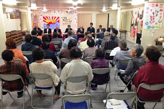 多目的室のような会場で、椅子に座った高齢の住民たちが前方のパネル席に座るスーツ姿の男性たちの話を聞いている写真