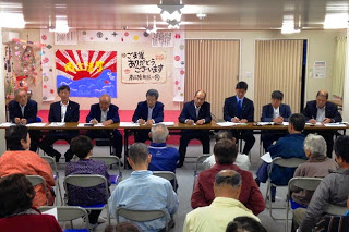 壁に「ご支援ありがとうございます」と書かれた用紙が掲示され、その前に長机を挟んで並んで座る関係者と、それに向かい合って座った地域住民が写っている写真