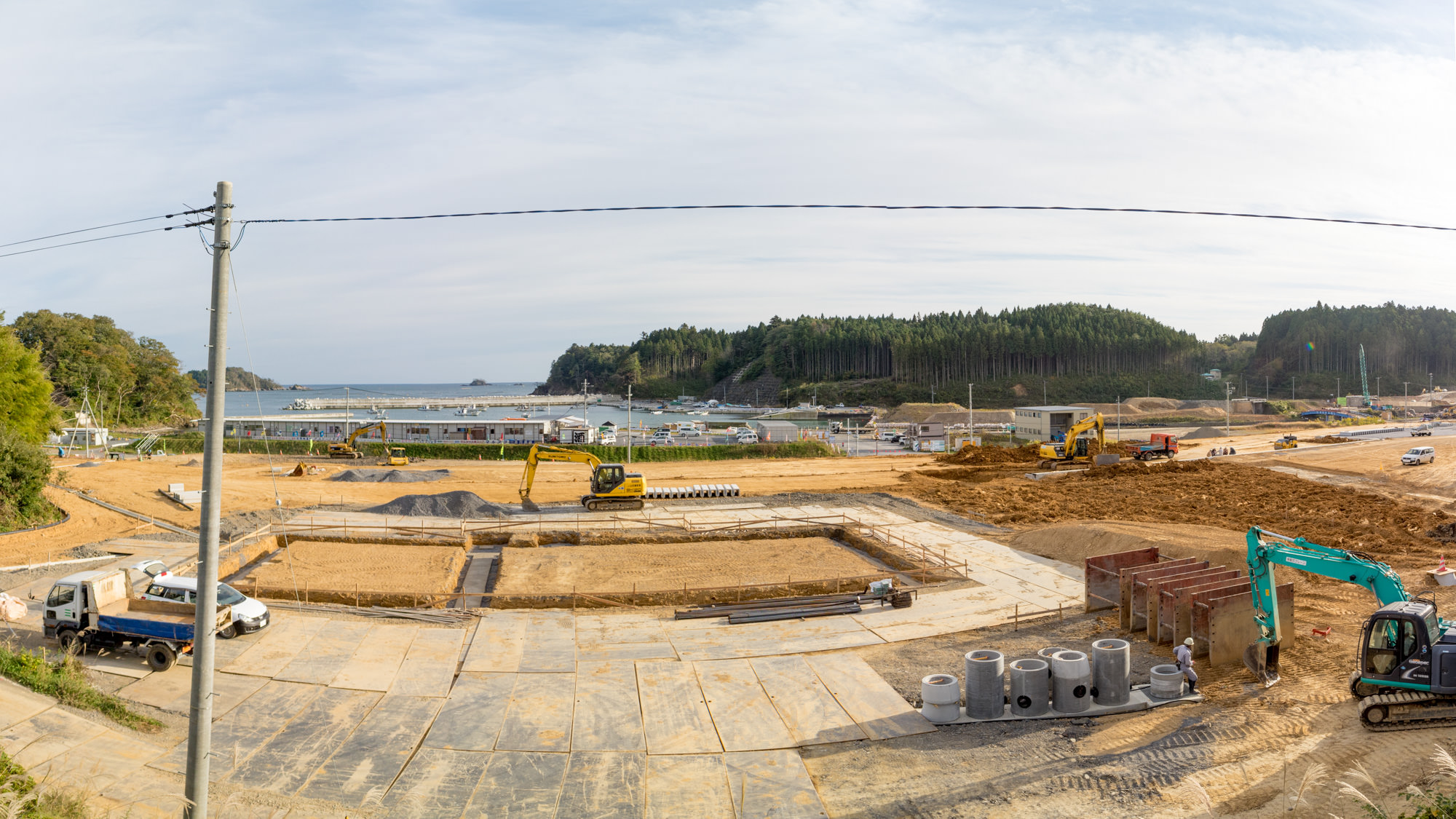 現場にはショベルカーやトラックなどの建設車両が複数稼働しており、地面は主に砂や土で覆われ、コンクリート製のパイプや資材も周囲に散在して、背景には水辺が広がり、マリーナや港に船が停泊している様子が見える海沿いの建設現場を写した写真