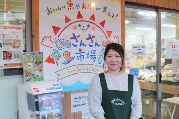 南三陸町の「さんさん市場」の看板の前で、緑色のエプロンを着た太田さんが笑顔で立っている写真