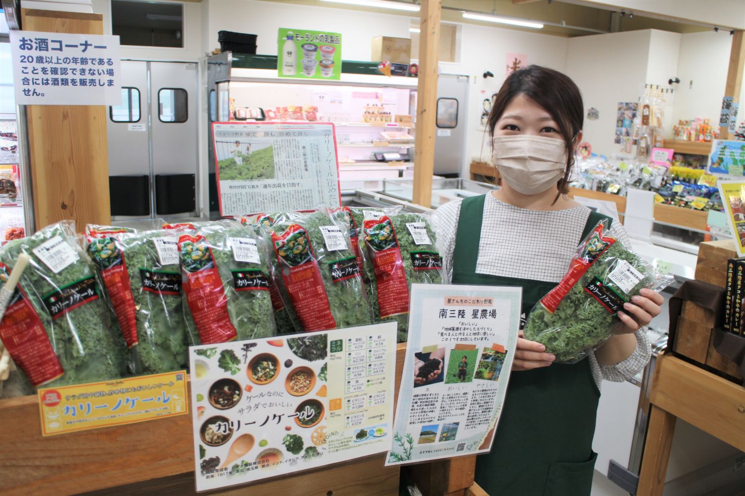 「さんさん市場」の青果コーナーで、太田さんが緑色のエプロンを着てカリーノケールを両手に持ち、おすすめ商品の前に立っている写真