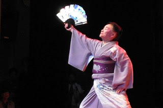 紫の着物姿の女性が舞台上で扇子を高く掲げて日本舞踊を踊っている写真