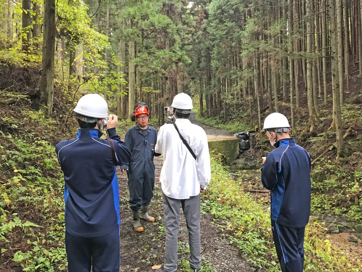 白いヘルメットをかぶった男性が赤いヘルメットの作業員を撮影し、その様子を高校生2名がそばで見ながらメモを取ったり写真を撮ったりしている森林の中の取材風景の写真
