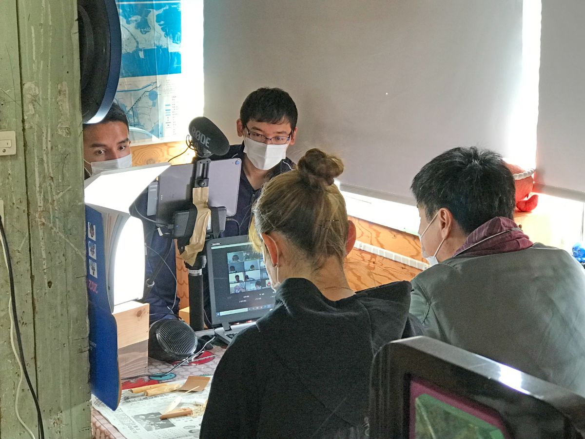 室内のテーブルに4名が向かい合って座り、マスクをつけた男女がカメラやタブレット、パソコンを使ってオンライン会議や撮影を行っている様子の写真