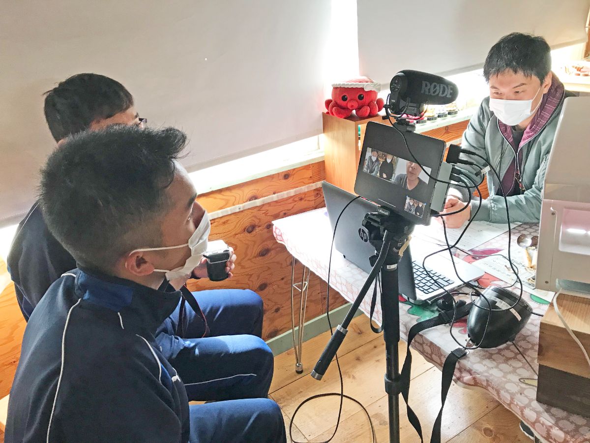 高校生の男子が手前に座り、机を挟んで関係者の男性が座り、マイクとカメラを使ってオンライン会議をしている三人の男性の様子を写した写真