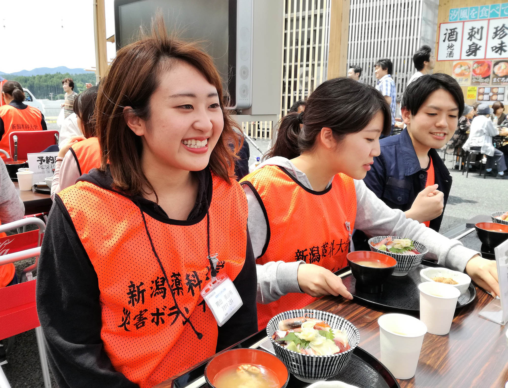 新潟薬科大学 災害ボランティア部』と書かれたオレンジ色のビブスを着た青山美沙望さんが、外の飲食スペースで友人と並んで座り、海鮮丼と味噌汁を前に笑顔で楽しんでいる写真