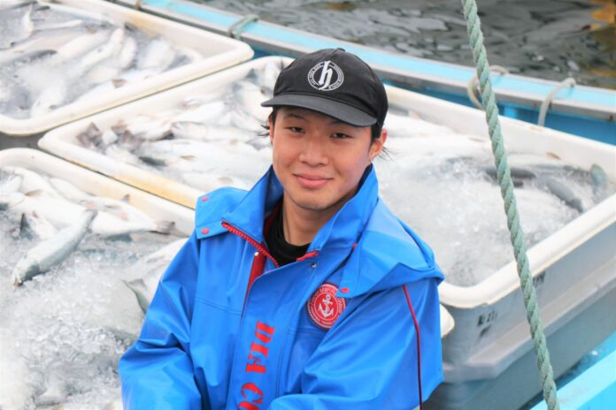 氷とともに入れられた多数の銀色の魚を背景に、青い漁業用合羽を着た吉岡さんが笑顔でカメラに向かって座っている様子の写真