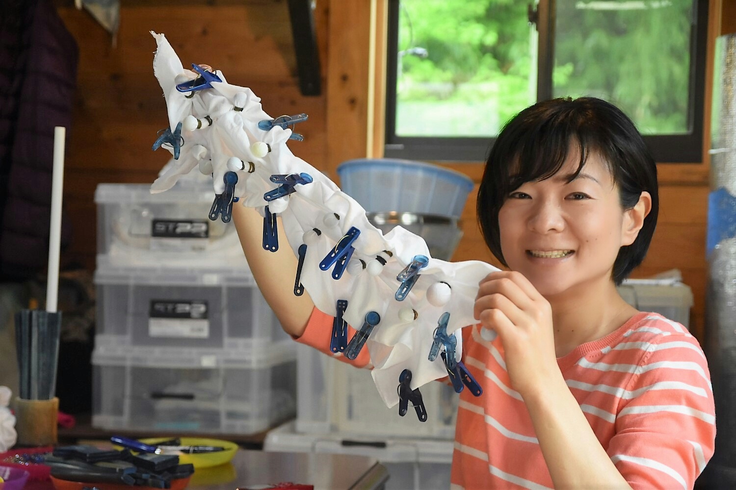 笑顔の女性が布に青い洗濯ばさみとゴムで結んだたくさんの白い玉を作り、絞り染めの準備を見せ、背景には収納ケースや道具が整然と置かれている藍染め作業中の写真
