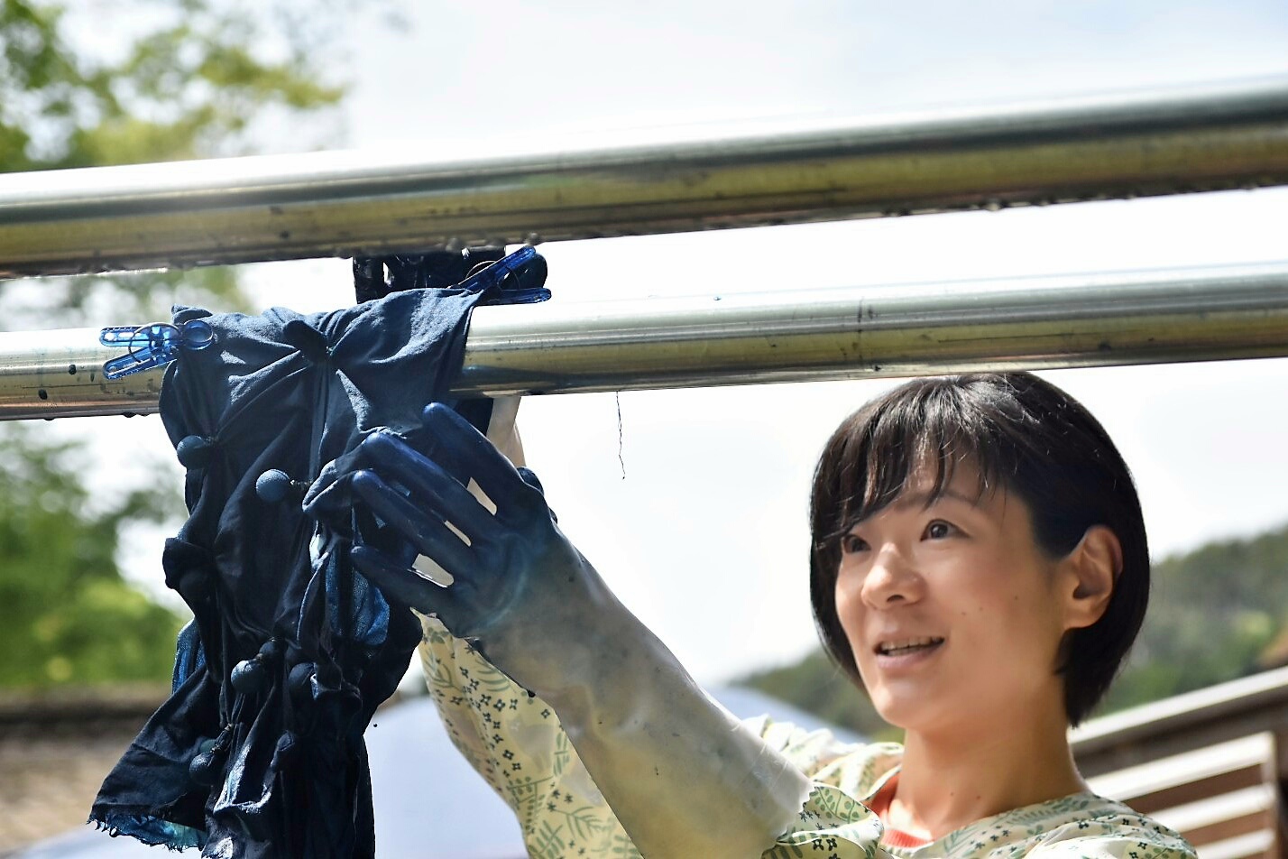 青い染料で染まった手袋をした女性が笑顔で、絞り染めの布を金属の物干し竿に干している写真