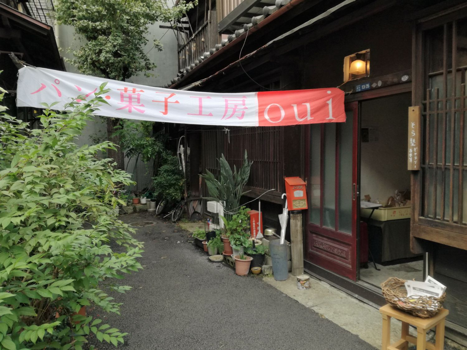 軒下には白と赤の横長の垂れ幕に「パン・菓子工房 Oui」と書かれ、細い路地の突き当たりに位置する古民家のパン屋の写真