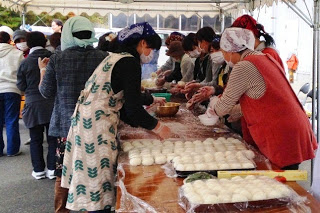 テントの下で、エプロン姿の女性たちが手際よく丸めた白いおにぎりを大量に並べて災害時の炊き出し準備をしている写真