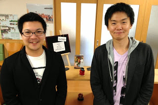 小さな黒板が飾られた室内の棚の前で、HAPPY南三陸部の2人の男性スタッフが並んで立っている写真