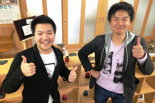 小さな黒板が飾られた室内の棚の前で、HAPPY南三陸部の2人の男性スタッフが笑顔で親指を立てて並んで立っている写真