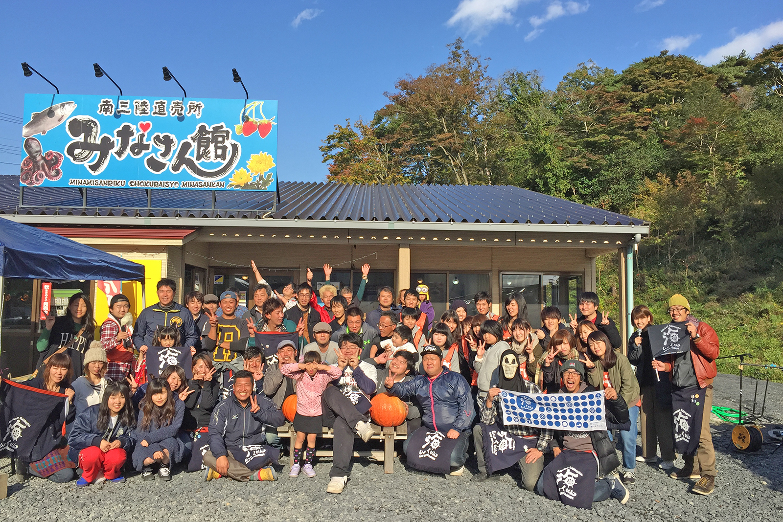 晴れた青空の下、「みなさん館」と書かれた看板のある建物の前で、老若男女の大勢の人々が笑顔で集まり、記念写真を撮っている活気に満ちた様子の写真