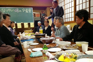 「なつかしい味の夕食会」と書かれた黒板を背にした和室で、高齢者を中心とする人々がにこやかに会話をしている写真