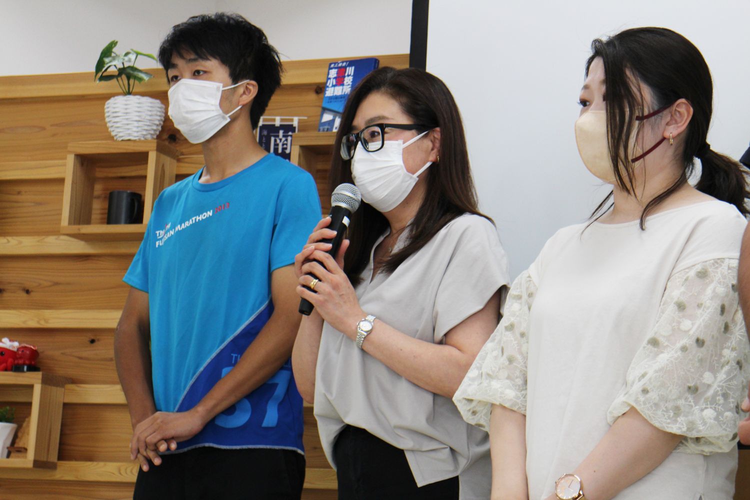 青いTシャツの男性、グレーの服の女性、白のブラウスを着た女性の3人がマスクを着けて並び、中央の女性がマイクを持って話している様子の写真