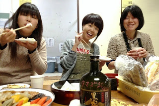 沢山の手作り料理や黒糖焼酎の瓶が置かれているテーブルの前に座った三人の若い女性が、和やかに食事を楽しみながら笑顔を見せている写真