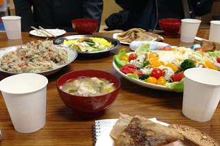 テーブルの上に並べられた色とりどりの料理と紙コップが写っており、奥には数人の人々が食事を楽しんでいる写真