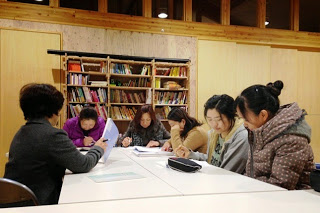 6人の女性が机を囲んで並んで座り、テキストを見たりノートを書いたりしながら日本語の勉強をしている写真