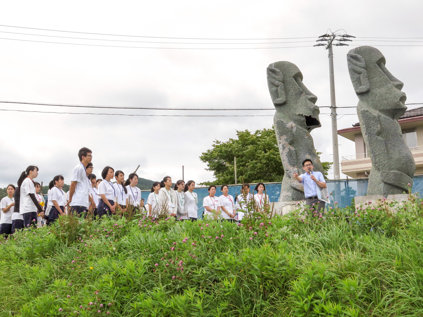 白いシャツ姿の高校生たちが草むらの前に立つ2体のモアイ像の前で男性の説明を真剣に聞いている写真