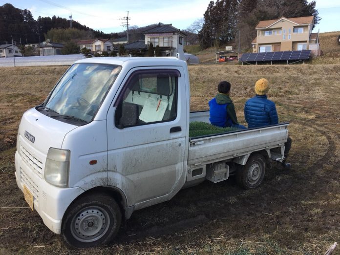 白い軽トラックの荷台に座っている2人の人物が、周囲の家々や太陽光パネルが並ぶ風景を静かに眺めている写真