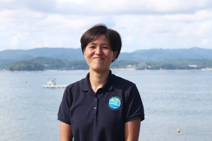 海を背景に、笑顔で直立する黒いポロシャツ姿の北島めいさんが写っており、後ろには船と山々が見える三陸の海辺で撮影された写真