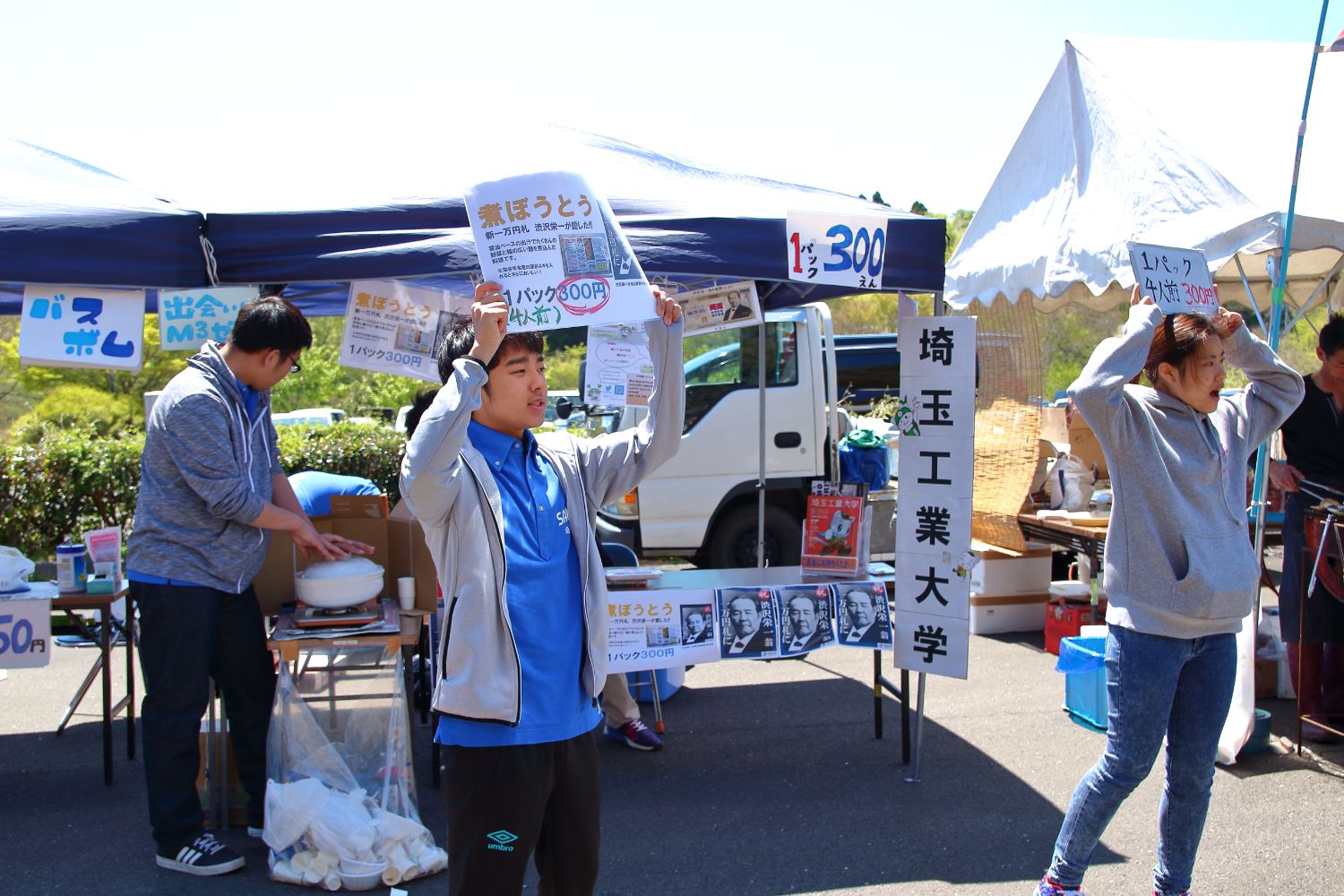 「埼玉工業大学」と書かれた看板が設置されたテントの前で、2人の大学生が手にチラシや看板を持ち上げながら声を上げ、呼び込みをしている様子の写真
