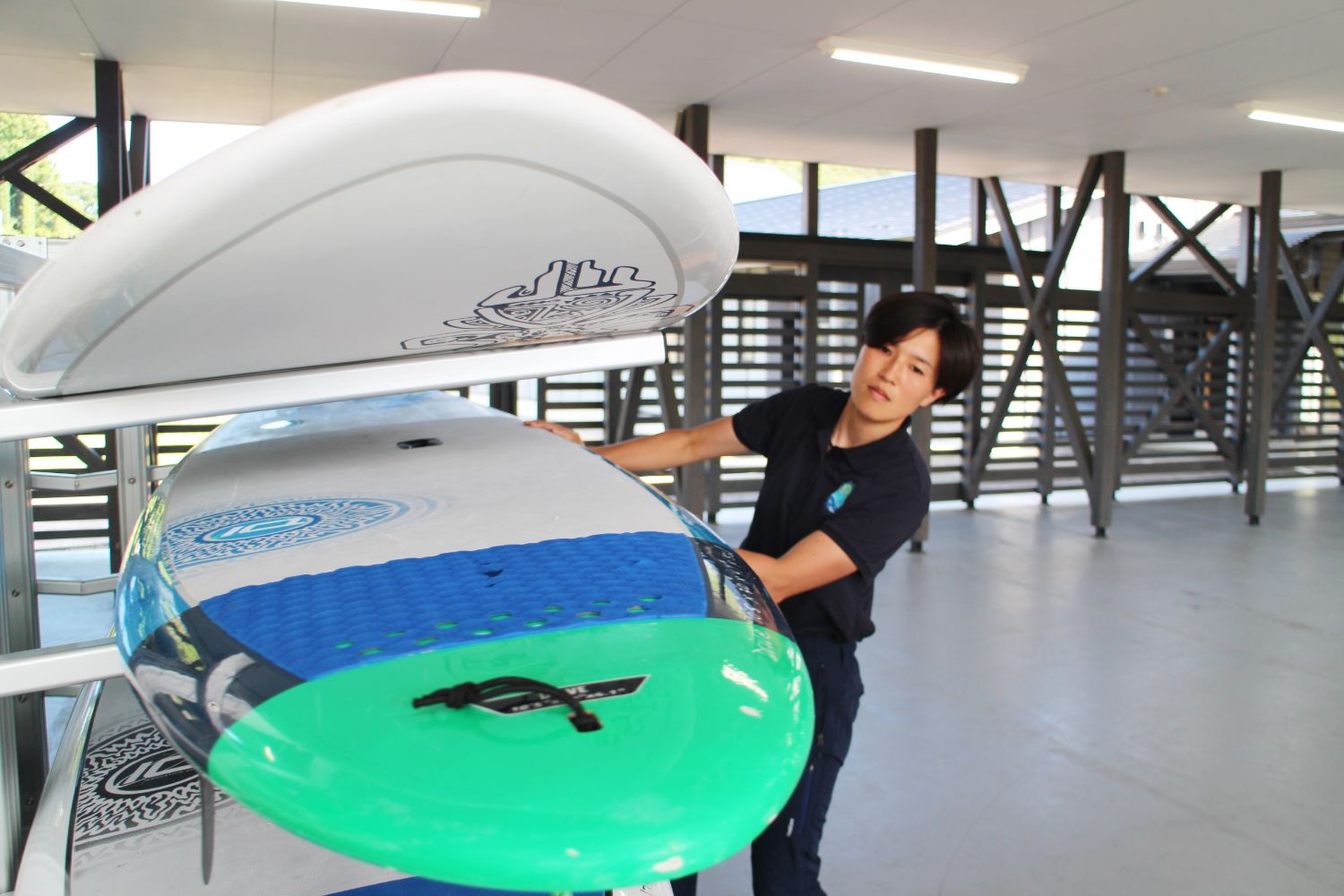 女性隊員がラックに収められた青と緑のスタンドアップパドルボードを取り出そうとしている様子が写っている写真