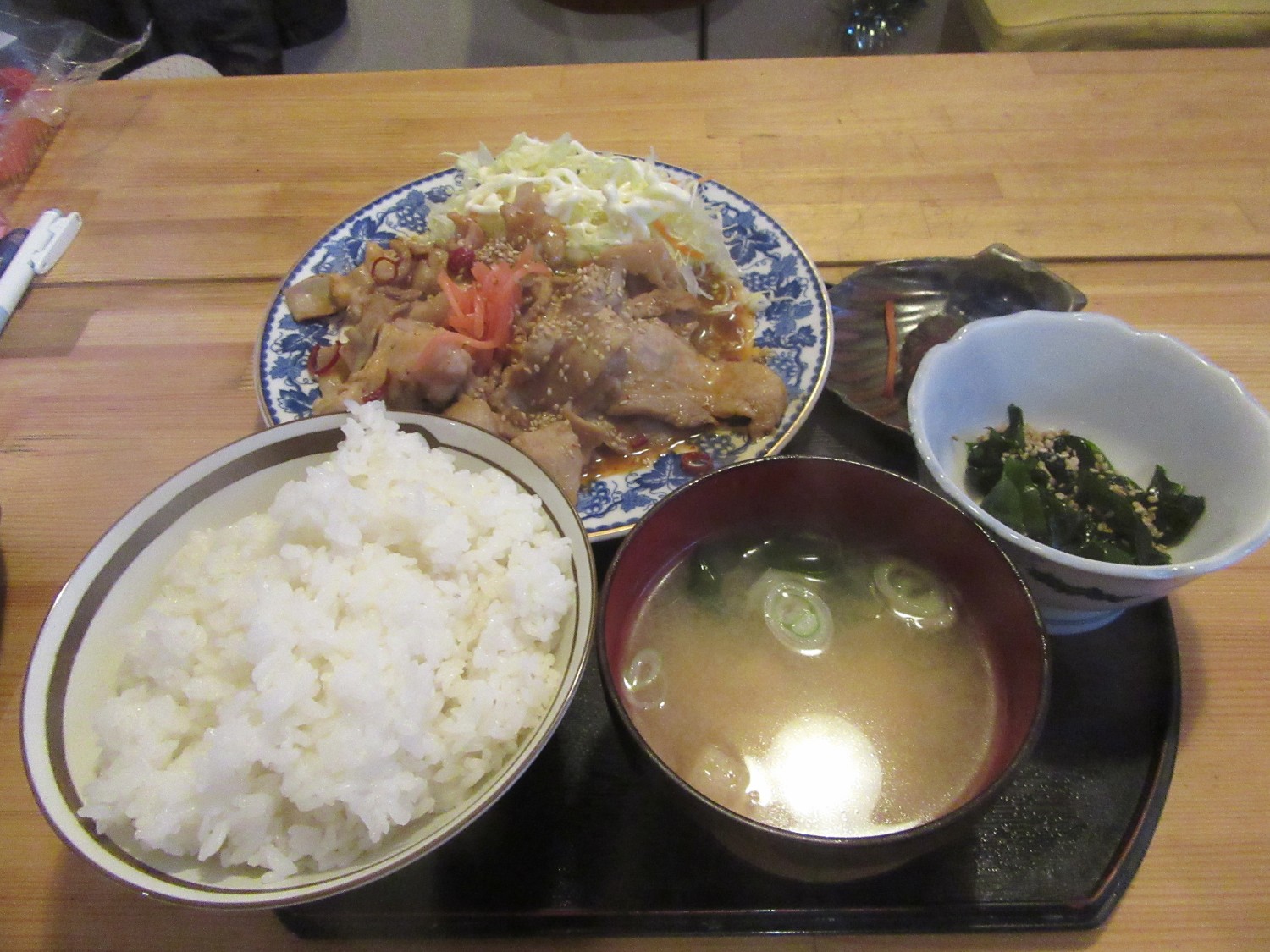 白ごはん、お漬物、味噌汁、小鉢、生姜焼きと千切りキャベツなどが盛られた和風定食がお盆に乗せられ木のテーブルに並べられている写真