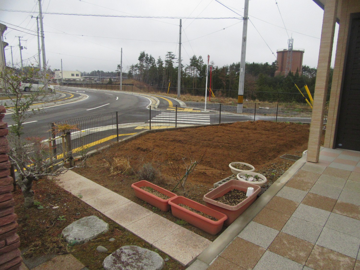 奥には小さな建物と林が見え、左側に向かってカーブする道路に面した家の前庭にプランターがいくつか置かれており、茶色く耕された土が広がっている写真