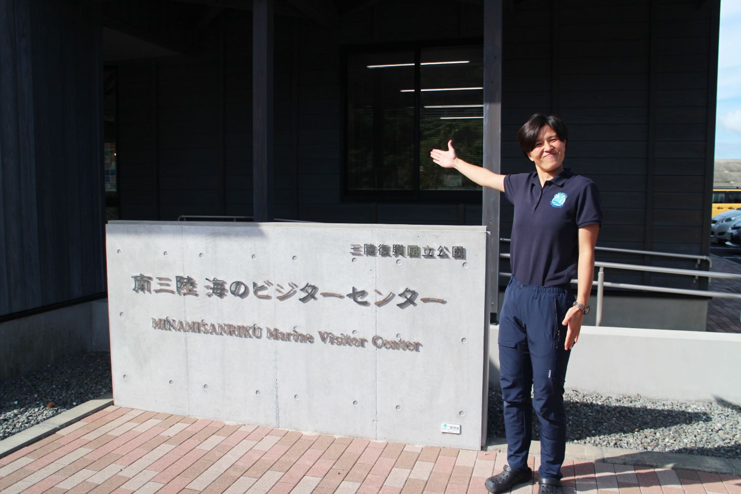 南三陸海のビジターセンターの前で、黒い制服姿の女性隊員が片手を広げて案内するように立っており、建物の看板とともに写っている写真