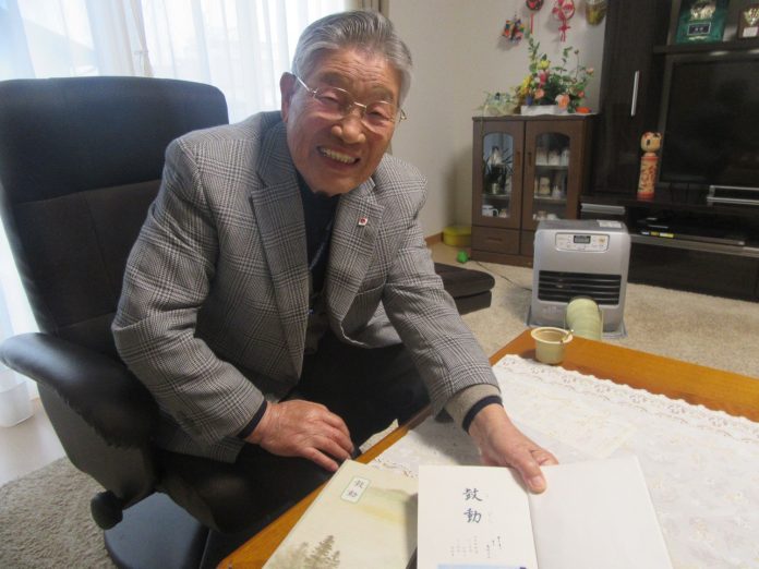 テレビや盾が飾られた棚のある室内で、椅子に座った笑顔のグレーのチェック柄のジャケットを着た高齢の男性が和室のテーブルの上で「鼓動」と書かれた句集を開いて見せている写真