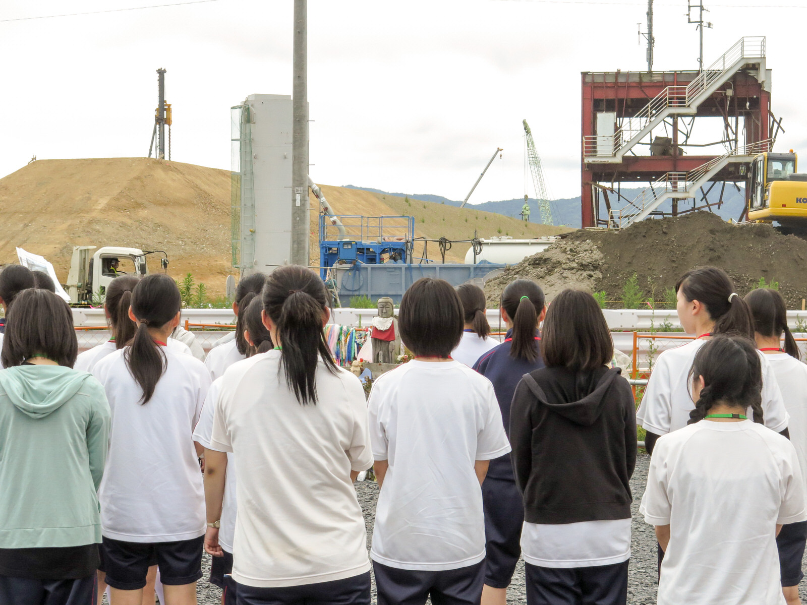 土の山や重機が見える復興工事現場の前で、ガードレール沿いに立つ小さな地蔵に向かって立っている高校生たちの後ろ姿の写真