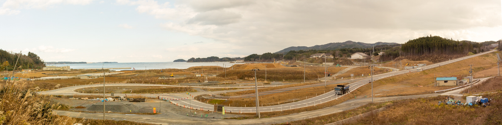曲がりくねった道路や一部の建設作業、数軒の建物が確認でき背景には山々が広がり、画像の左側には海が見える海沿いの地域をパノラマで写した写真