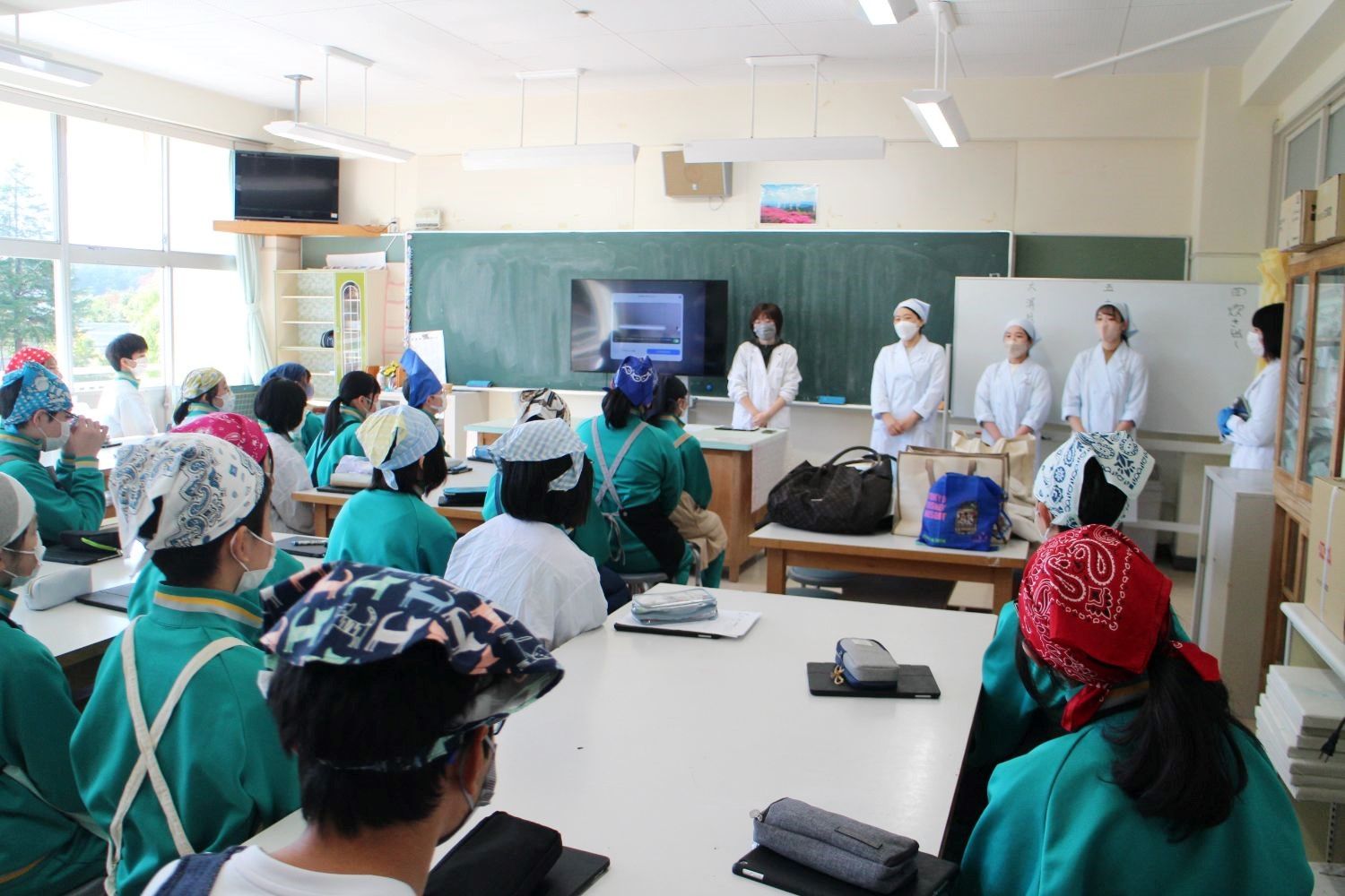 教室で三角巾と緑色のジャージを着た生徒たちが前方の黒板の前に置かれたモニターとその横に並んで立った白衣姿の4人の発表者を見ている写真